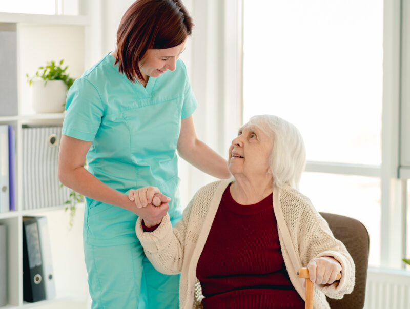 Old,Lady,Leaning,On,Nurse,Hand,To,Stand,From,Chair